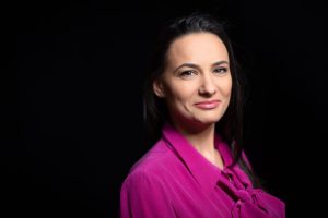 Teodora Ghenciu zâmbește, purtând o bluză roz, pe un fundal întunecat.