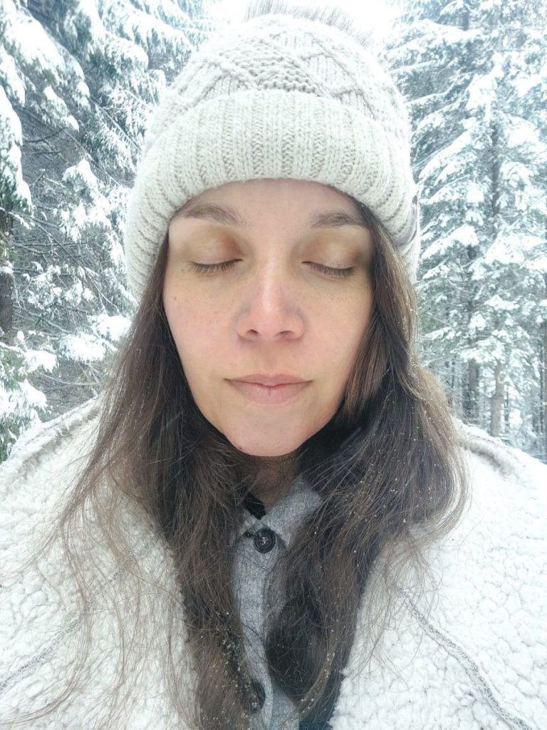 Woman with closed eyes, wearing a hat, in a snow-covered forest.