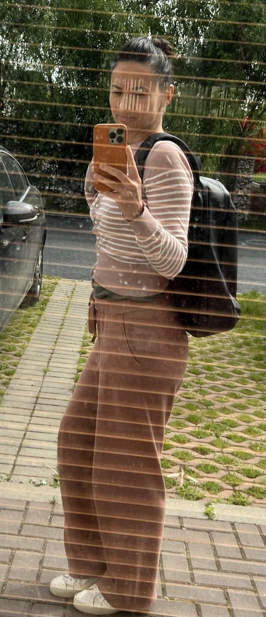 A woman takes a selfie in a mirror, wearing a backpack and casual clothes, with an urban landscape in the background.