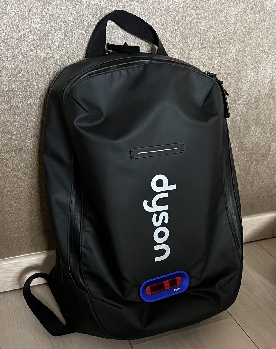 Black backpack with an air pollution measuring device, against a wall background.