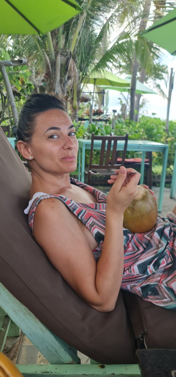 Woman relaxing on a lounge chair, holding a coconut, surrounded by palm trees.