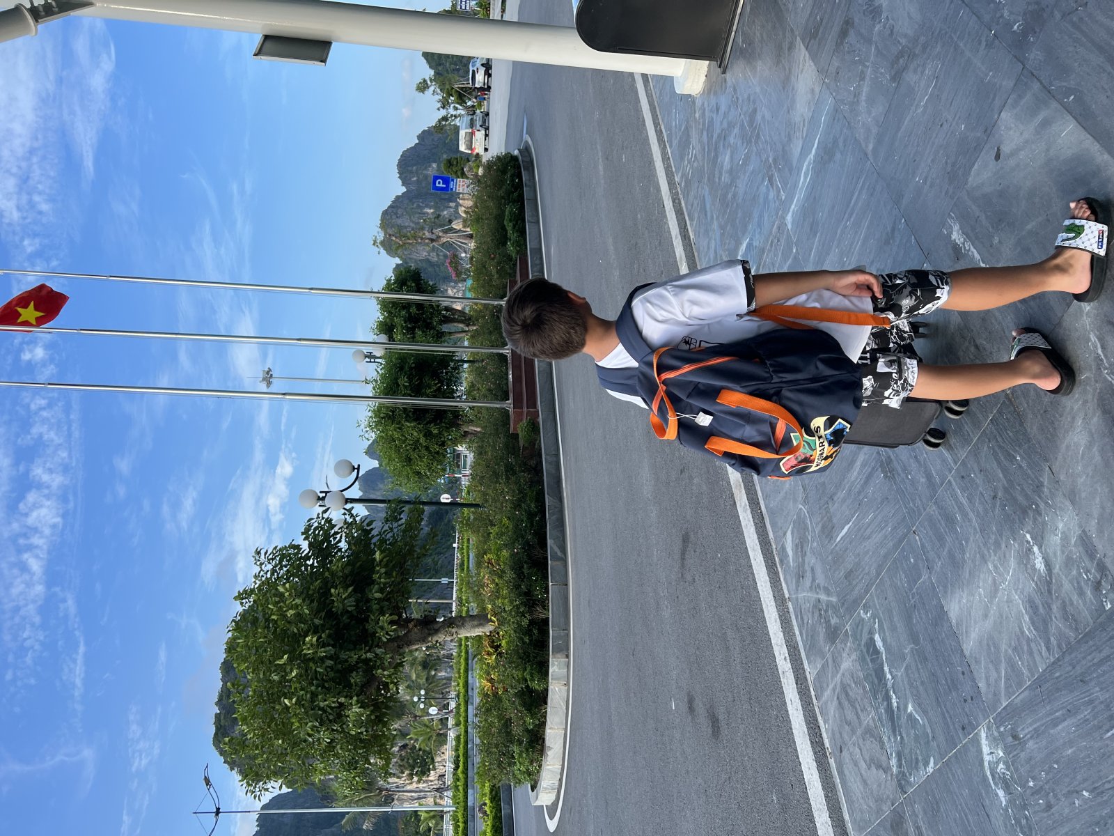 A boy with a backpack walks towards an urban landscape in Vietnam, featuring flags and mountains in the background.