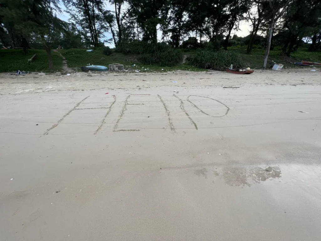 Message written in the sand on a beach, with debris and vegetation in the background.