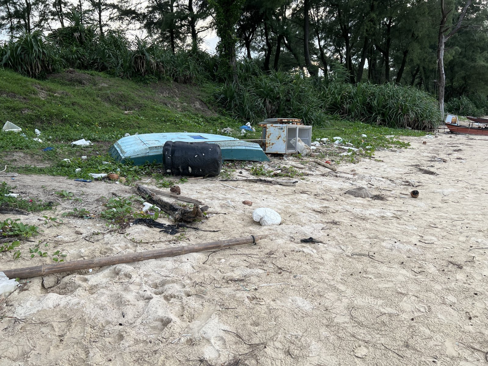 Dirty beach littered with plastic and debris, including an abandoned boat and vegetation in the background.