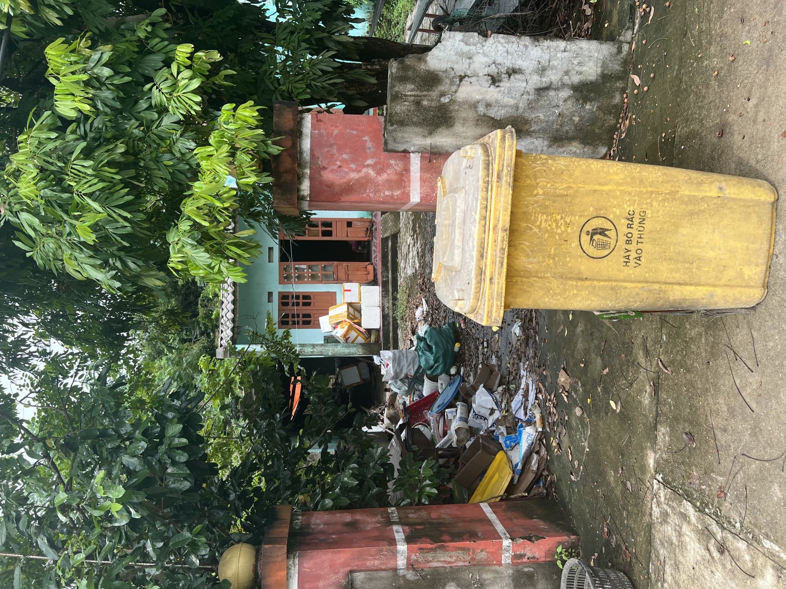 A yellow trash bin in a lush area with visible waste and greenery.