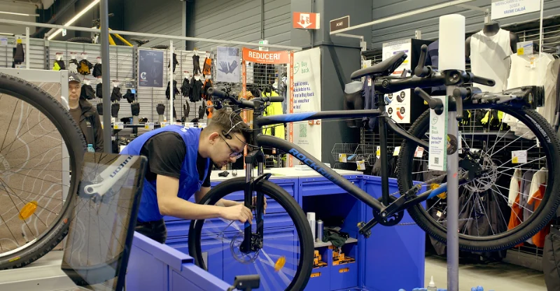 Un angajat repară o bicicletă într-un atelier Decathlon, cu diverse produse sportive în fundal.