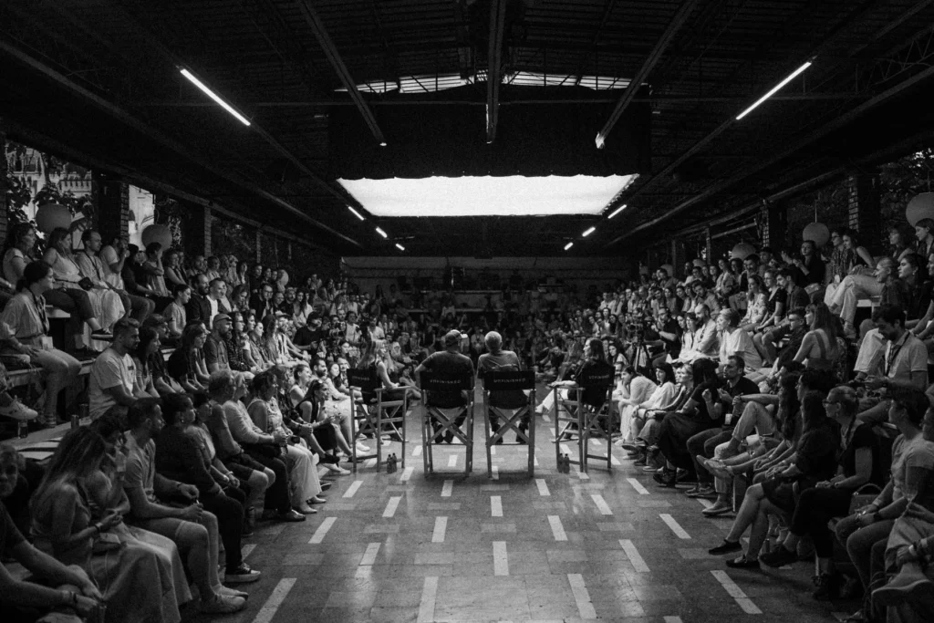 A packed hall of spectators listening to a discussion panel. Vibrant atmosphere and engagement.