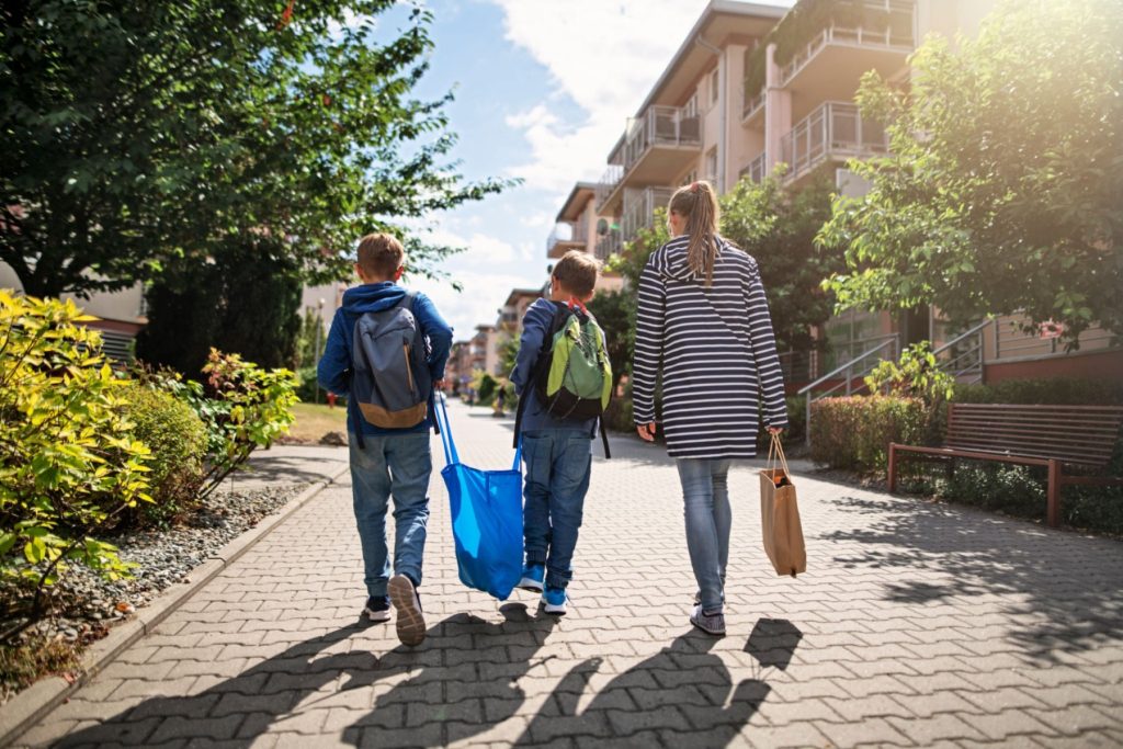 Three people, two children and a woman, walk along a pathway in a neighborhood, carrying backpacks and bags.