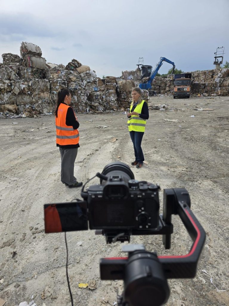 Two women discuss in a recycling facility, with machinery and waste piles in the background.